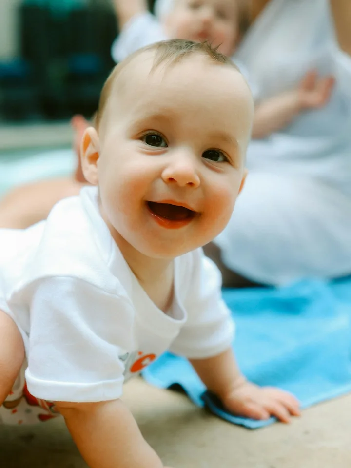 A baby in a white one-piece smiling, seated on a plain studio backdrop