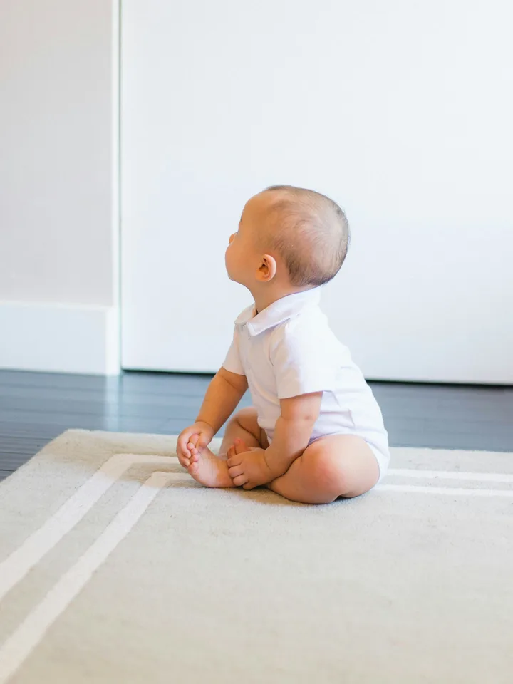 A baby in a white bodysuit sitting on a wooden floor