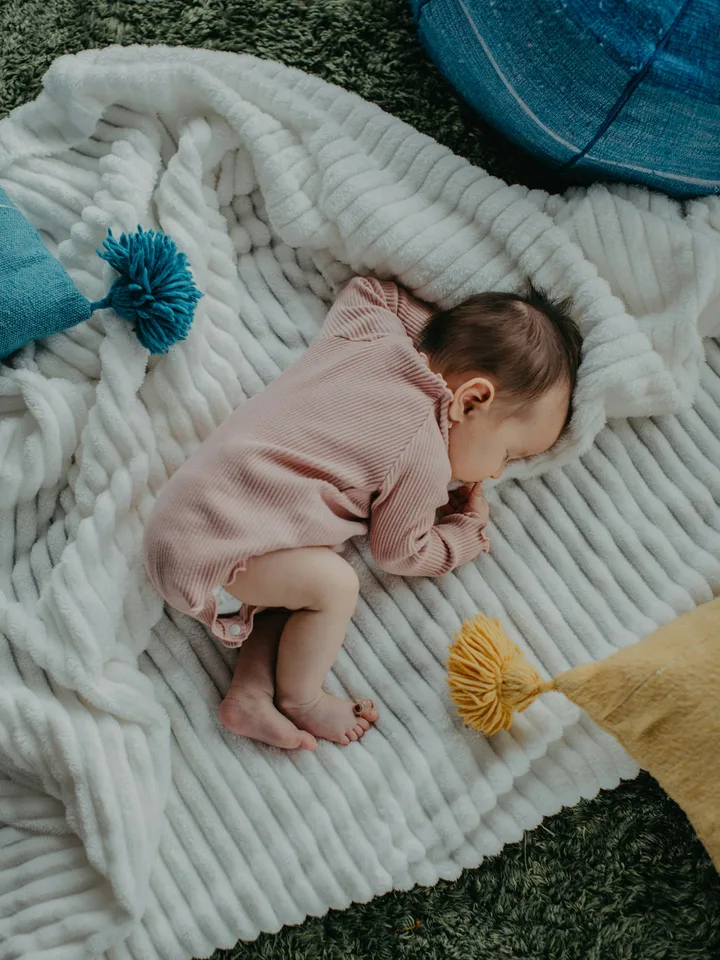 A baby asleep on a white bed, arms up beside the head