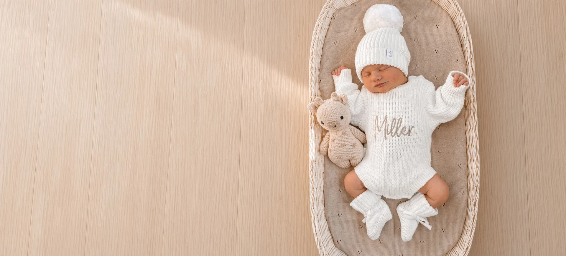 A sleeping baby in a white knitted romper and pom-pom hat, lying in a wicker basket beside a knitted bunny
