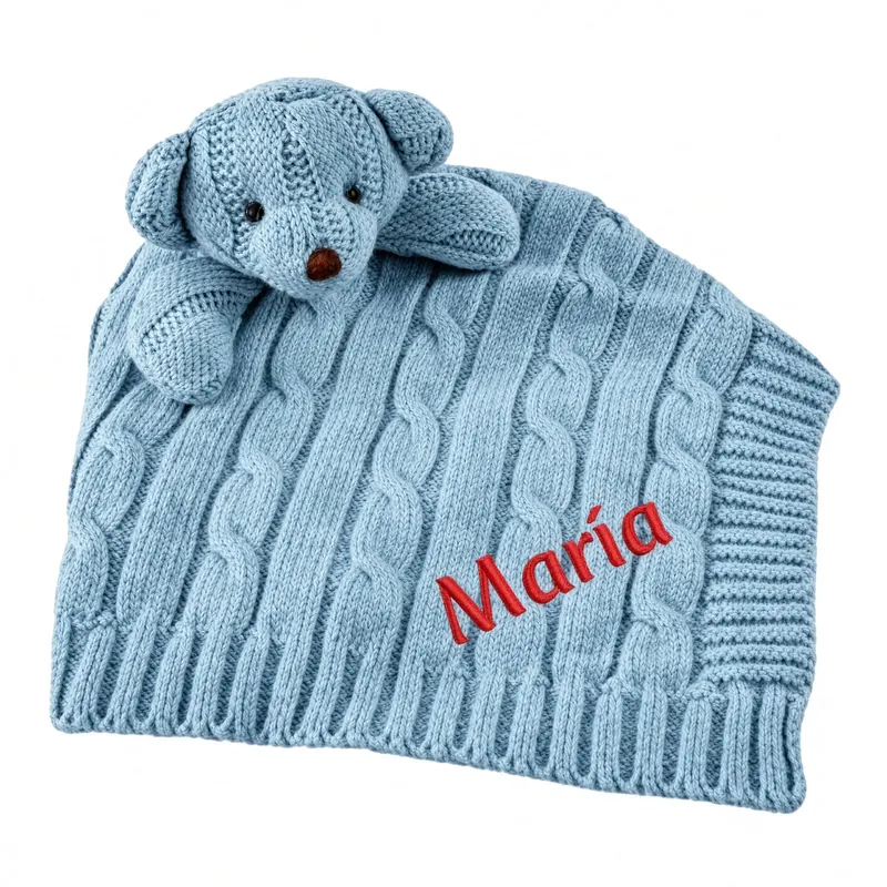 Light blue cable-knit baby blanket topped with a knitted teddy head and a name embroidered in red