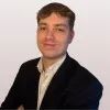 Man in a dark suit jacket and a white shirt against a plain light wall