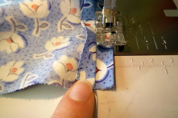 Blue floral cloth feeding under a presser foot, a thumb guiding the seam