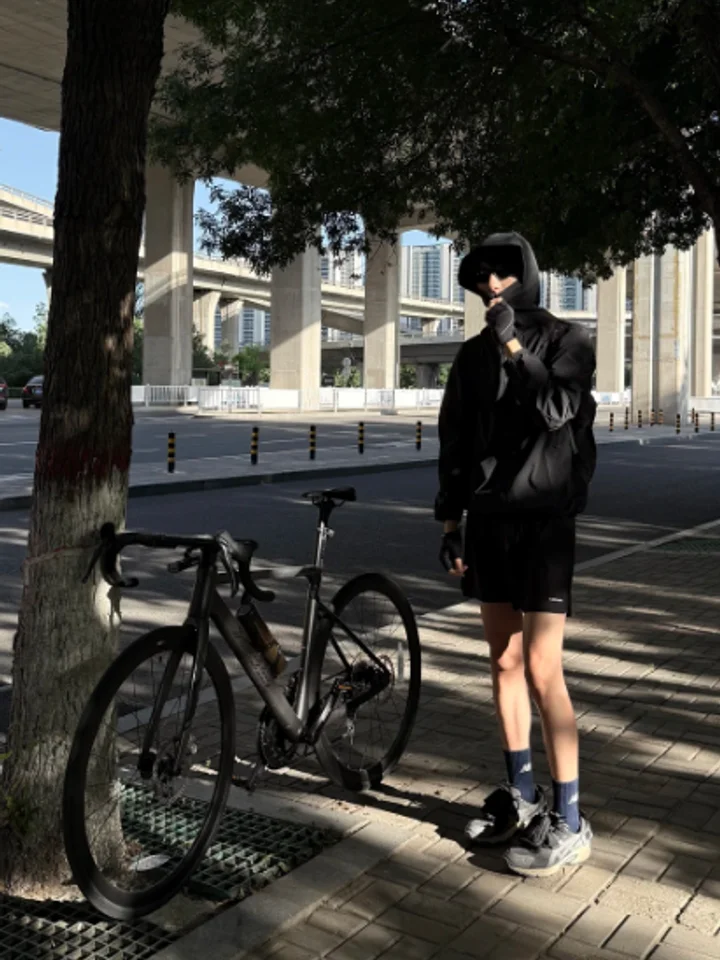 Standing beside a road bike on a city street, dark jacket and black shorts in the shade of a tree