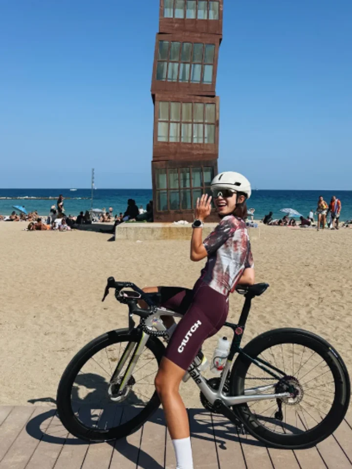 Leaning on a road bike on the sand with one hand raised, a beach and a tower of windows behind