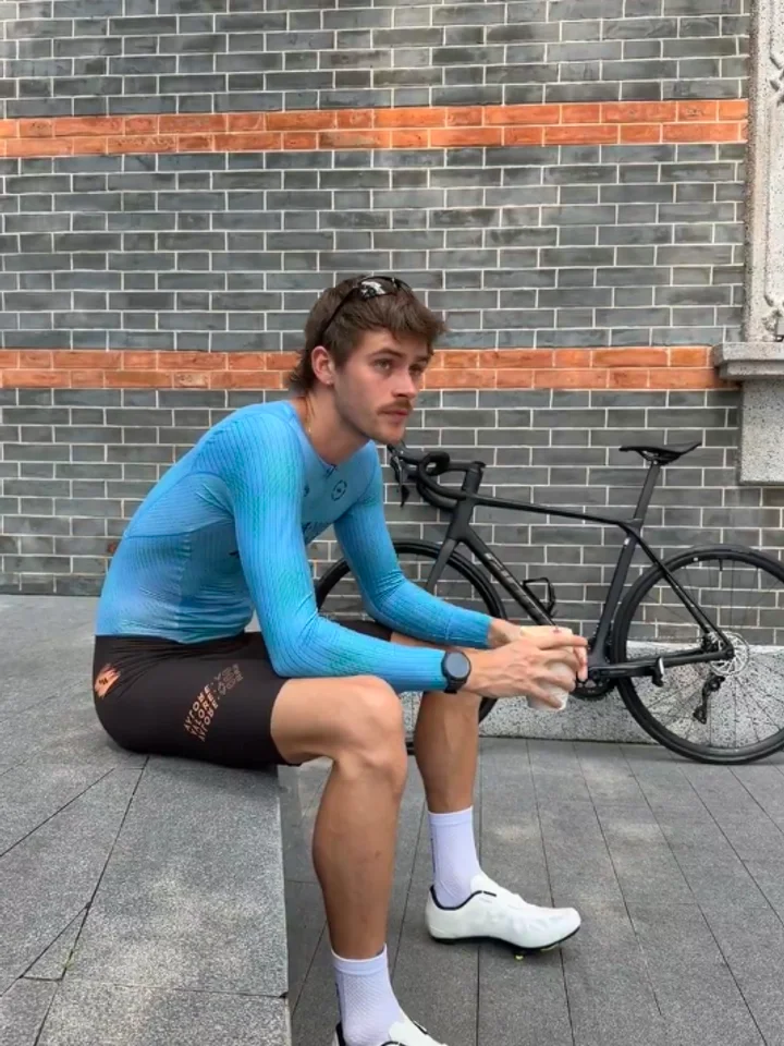 Seated on a step against a tiled wall, blue long sleeves and black bibs, a bike propped alongside