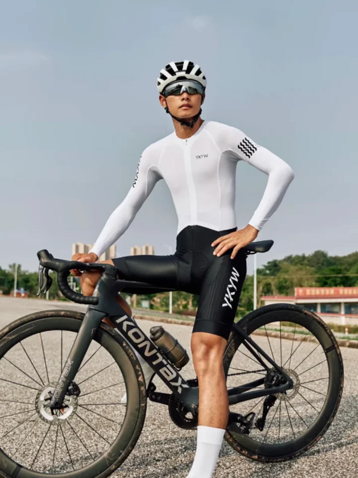 Holding a road bike at rest in white long sleeves, a striped helmet on and the bars in one hand