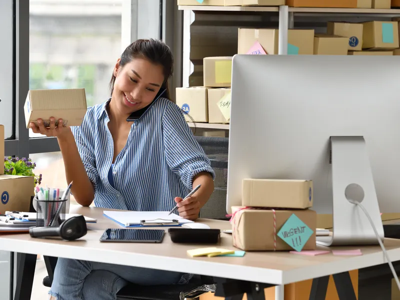 An online store owner on the phone at her desk, surrounded by cardboard shipping boxes — an e-commerce seller preparing a reorder