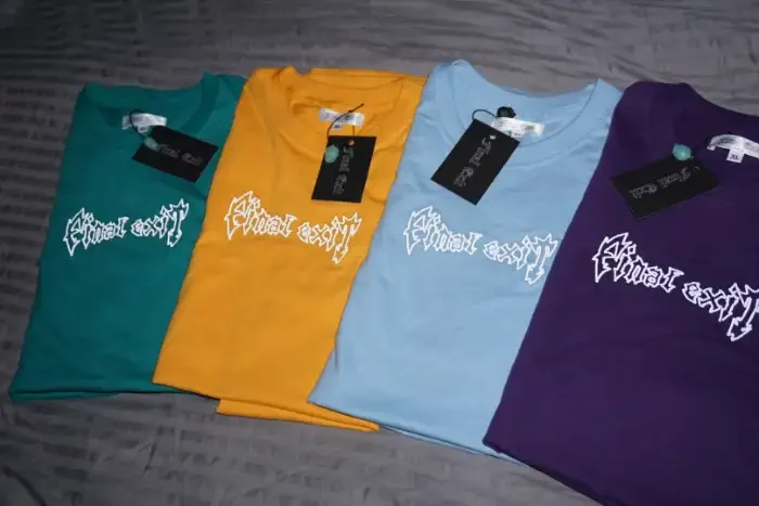 Four folded tees in green, yellow, blue and purple carrying the same stitched wordmark