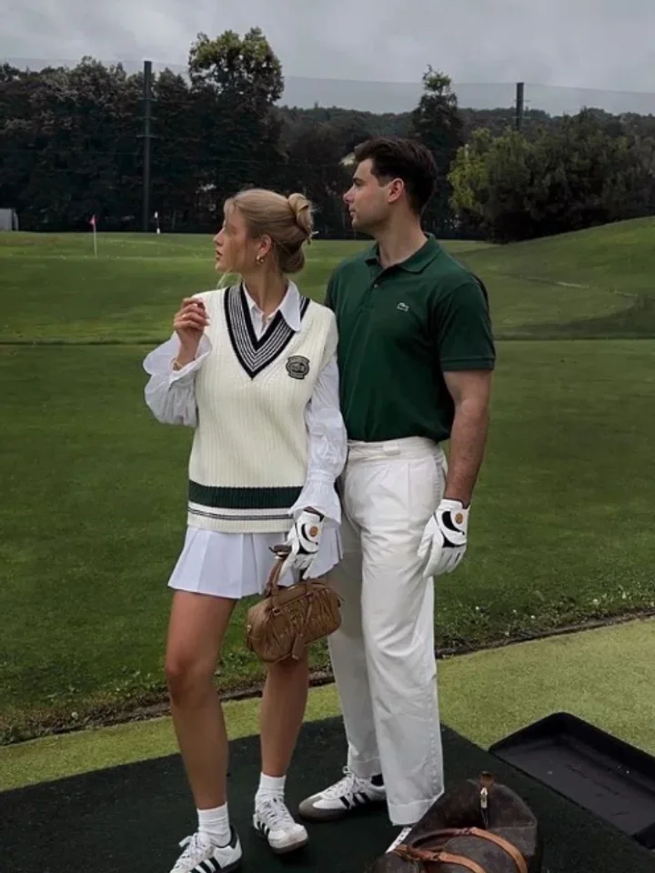 Standing on a black tee mat in front of a treelined fairway, a white sweater vest with green trim beside a dark green polo and white trousers