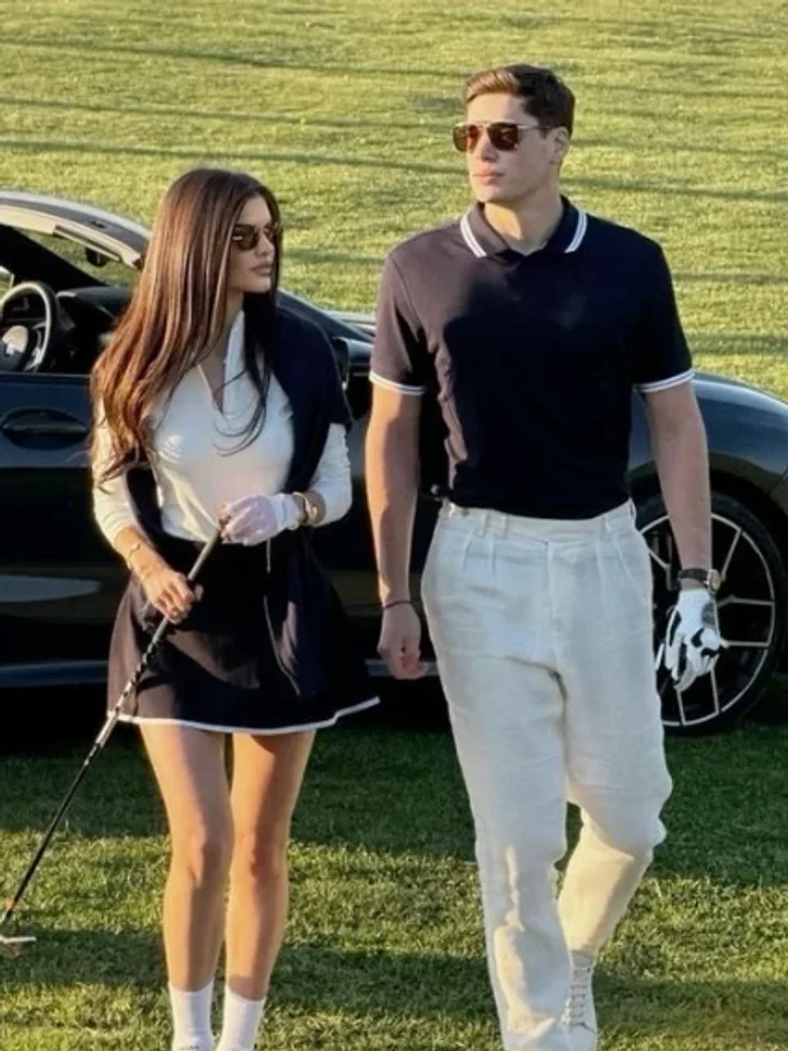 Walking past a black sports car on the grass, a white top with a sweater over the shoulders beside a black polo and cream trousers