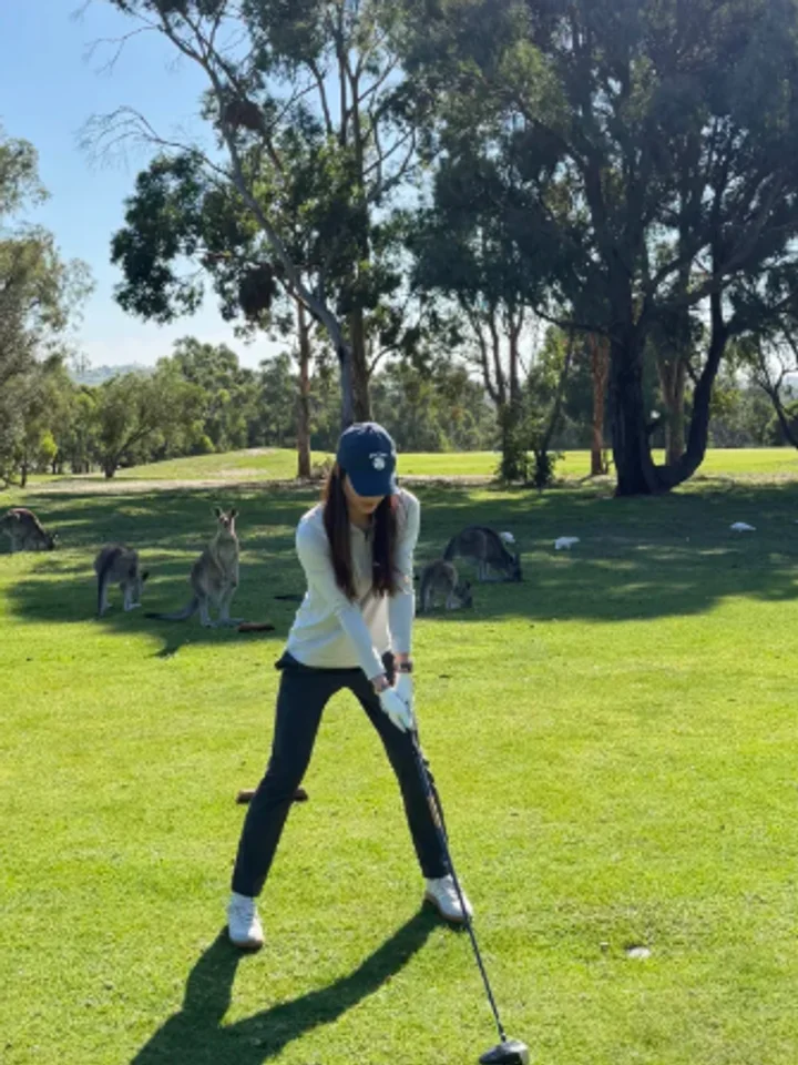 Swinging a driver on a fairway with kangaroos grazing behind, a navy cap over a white long-sleeve top and dark trousers