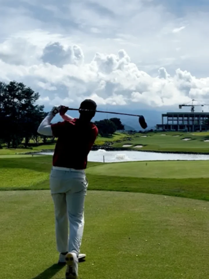 Viewed from behind mid-backswing over a lake and bunkers, a burgundy polo with white trim and cream trousers
