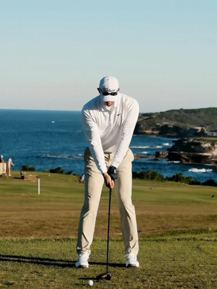 Addressing the ball on a clifftop tee above the sea, a white cap and long-sleeve top over stone-coloured trousers