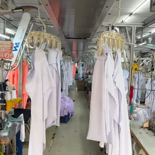 Moving along an overhead rail, white and pale lavender garments on hangers above a factory aisle.
