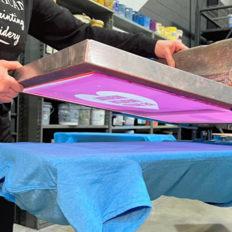 Mesh screen lifted off a blue tee on the press bed, the white design still sitting on the stencil