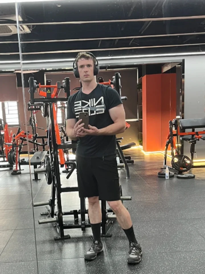 Mirror selfie in a black graphic tee and shorts, headphones round the neck, orange-framed machines behind