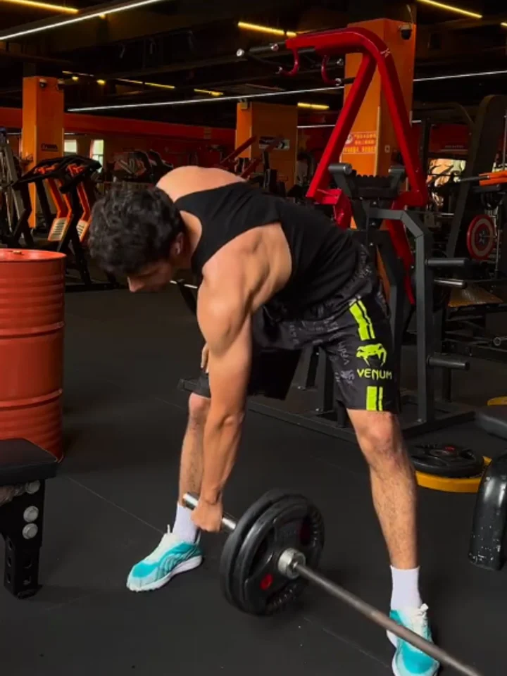 Lifting a loaded barbell off the floor in a black tank and panelled shorts, head down beside an orange barrel