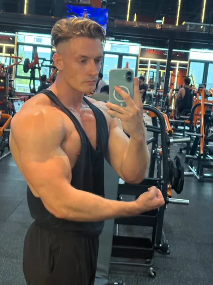Flexing side-on in a black lifting singlet, the phone raised in the other hand against a mirrored wall