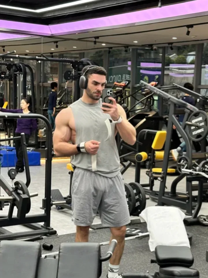 Standing in a grey sleeveless tee and shorts for a mirror selfie under a purple ceiling light