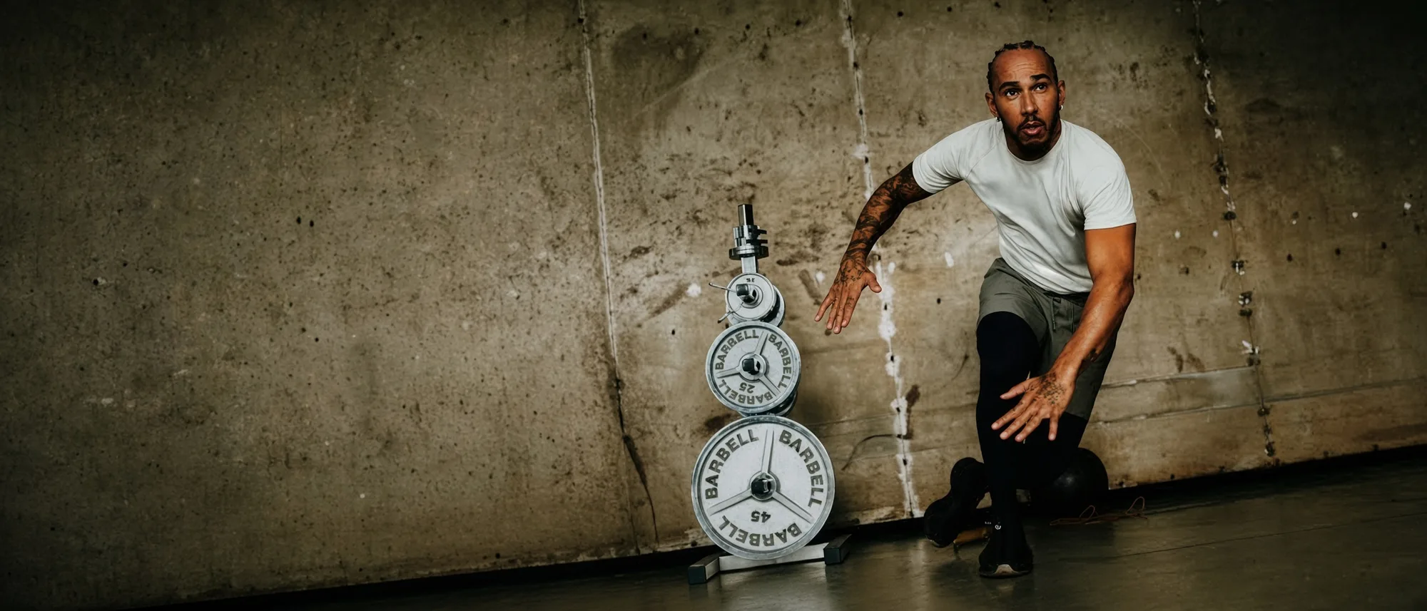 Man in a white tee and shorts crouched with one hand reaching out beside a stack of barbell plates on a concrete wall