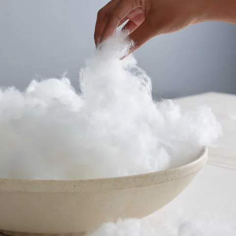 Hand lifting a tuft of white down-alternative fill from a stoneware bowl