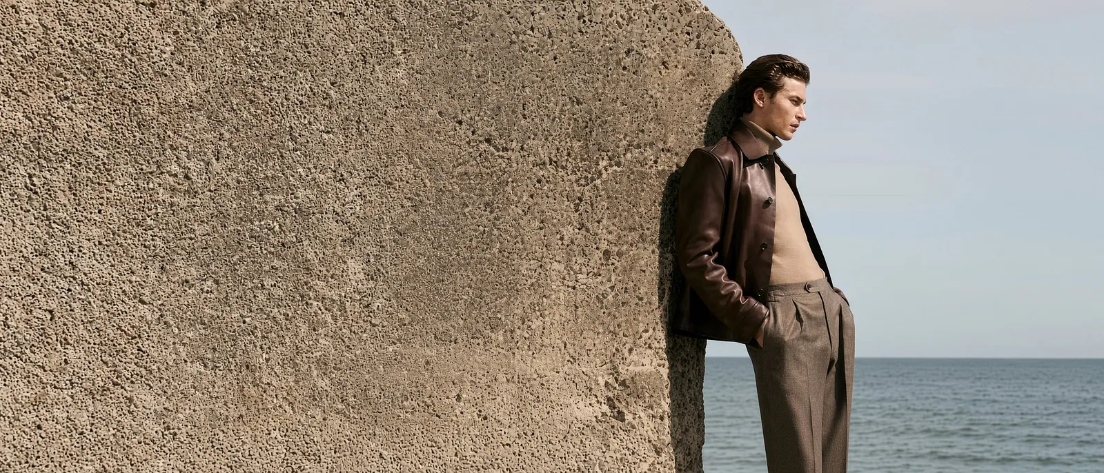 Custom brown leather jacket and pleated trousers on a model leaning against a concrete wall by the sea