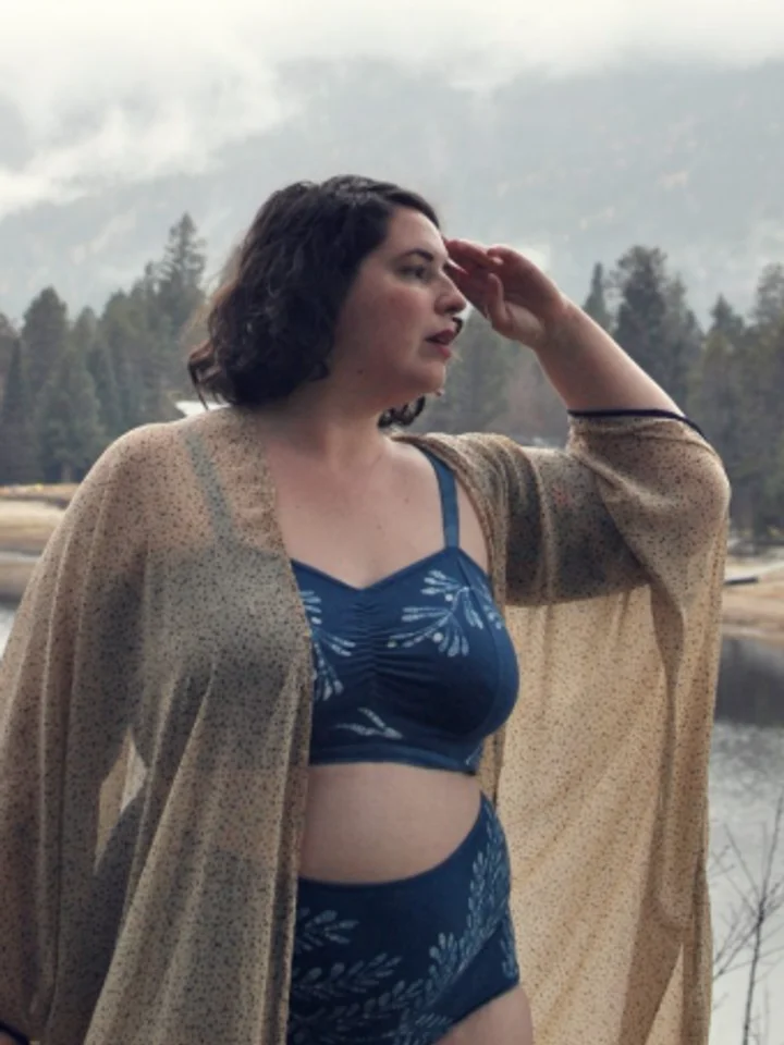 A leaf-print bra and high-waist brief layered under an open beige cover-up, worn on a lake shore