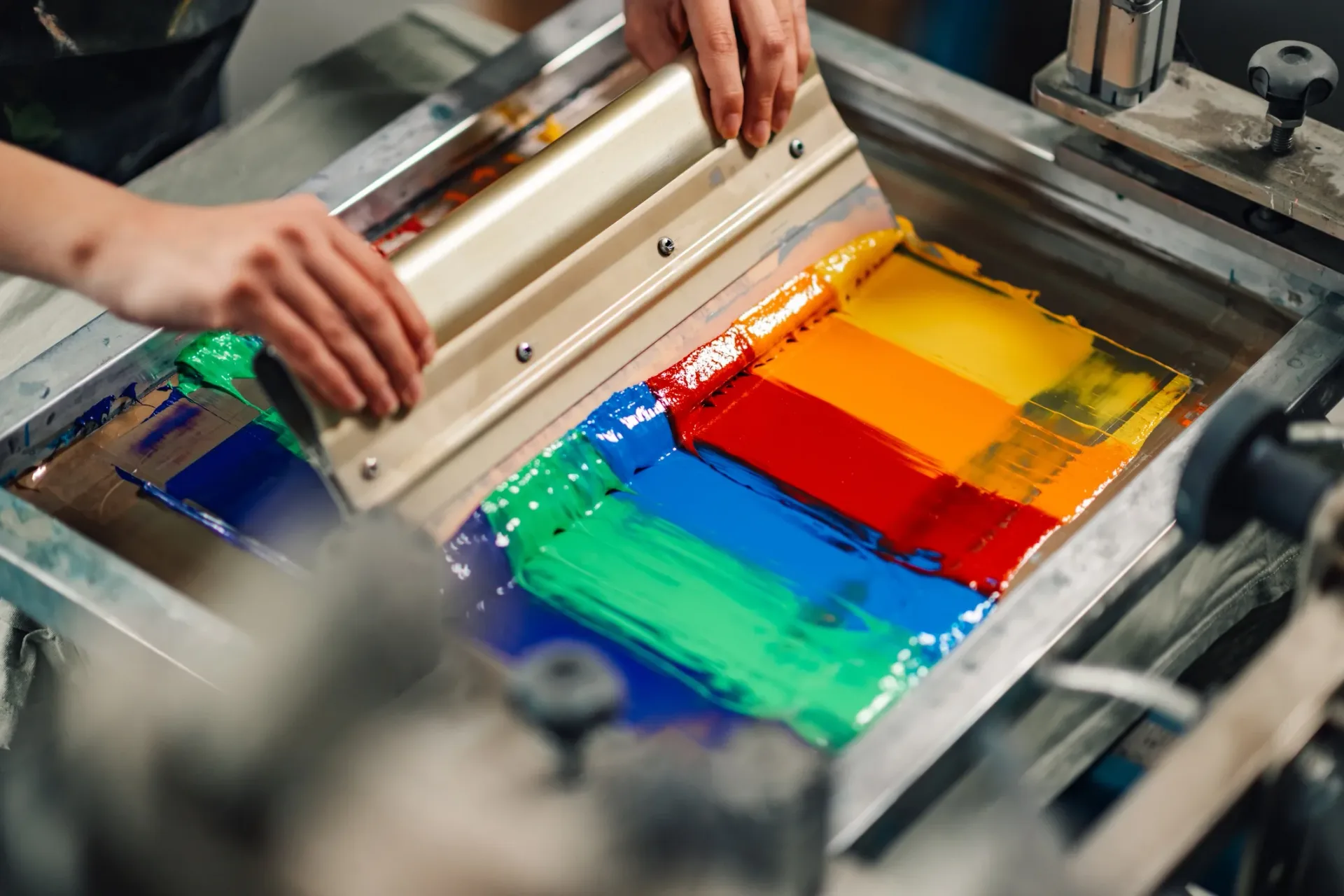 Hands pulling a squeegee across a screen flooded with four spot colours