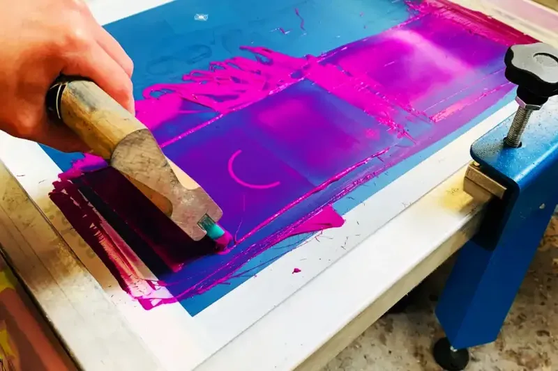 Hands pulling a squeegee through a bead of magenta ink across a mesh screen on a press