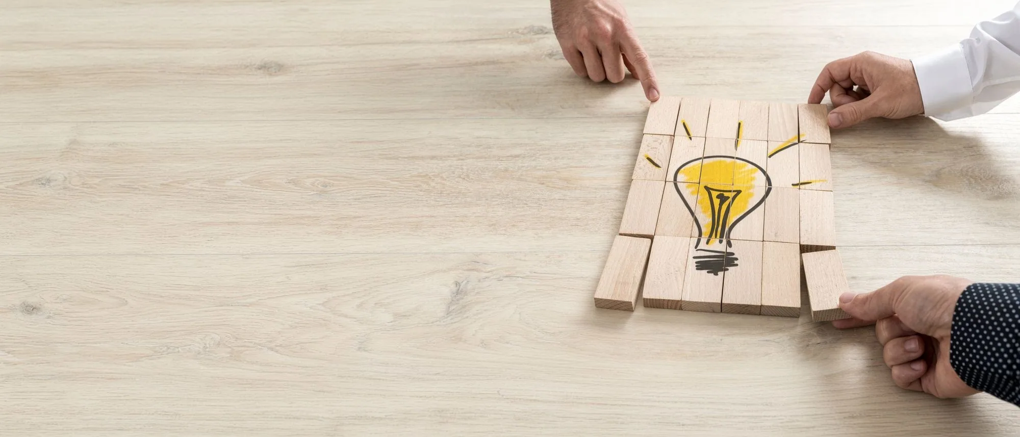 Two pairs of hands fitting wood blocks around a drawn lightbulb on a pale desk