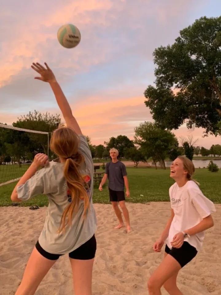 A woman playing beach volleyball in a graphic tee and shorts at sunset