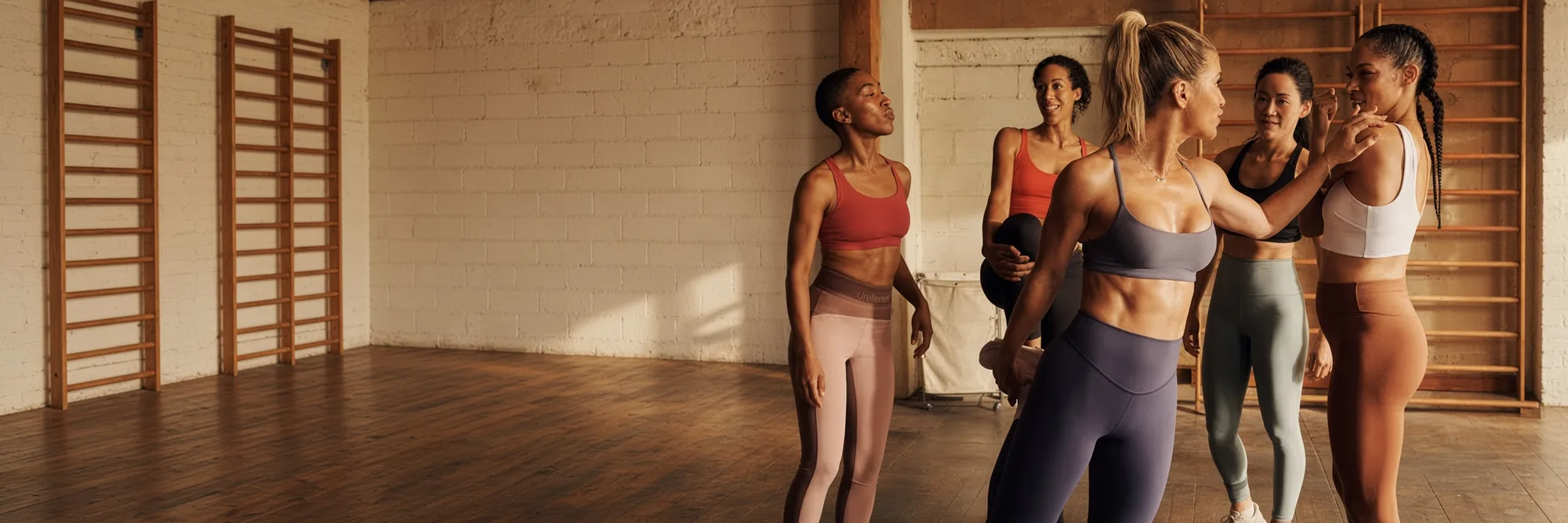 Five women in sports bras talking together in a studio, wooden wall bars along the wall behind them