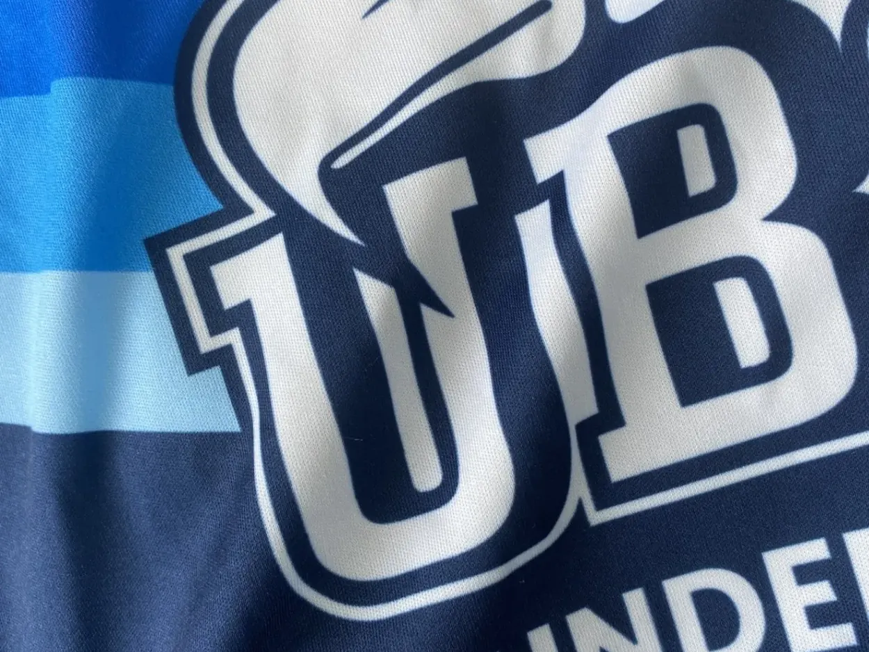 Close-up of a dyed polyester jersey — navy ground, light-blue stripes and a team crest, all of it colour sitting in the cloth rather than on it