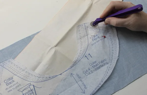 A rotary cutter following a marked paper pattern laid over blue fabric