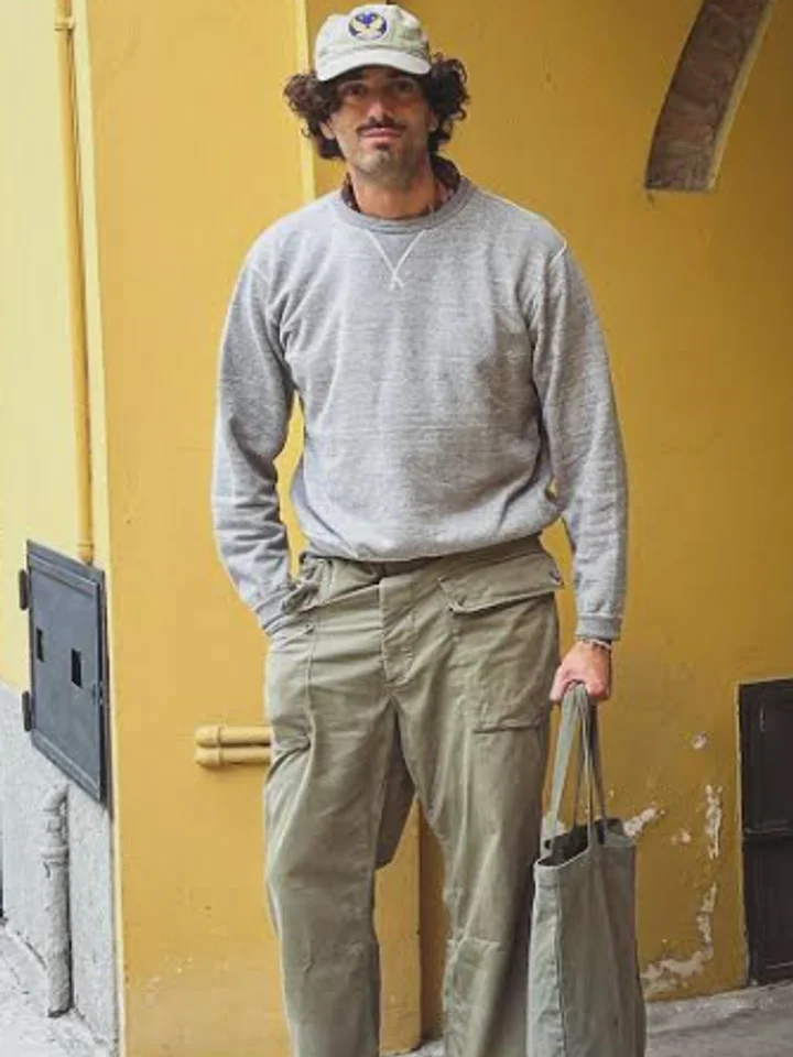 A customer in a grey crewneck sweatshirt and khaki cargo trousers, wearing a cap and carrying a tote, against a yellow wall