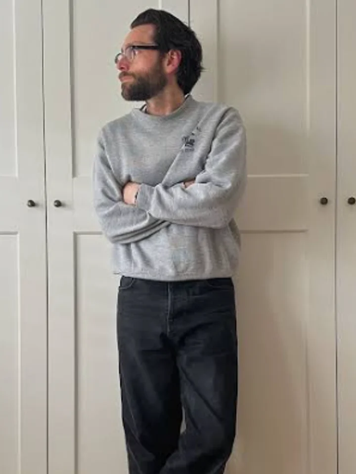 A customer in a grey crewneck sweatshirt standing with arms crossed beside white panelled doors
