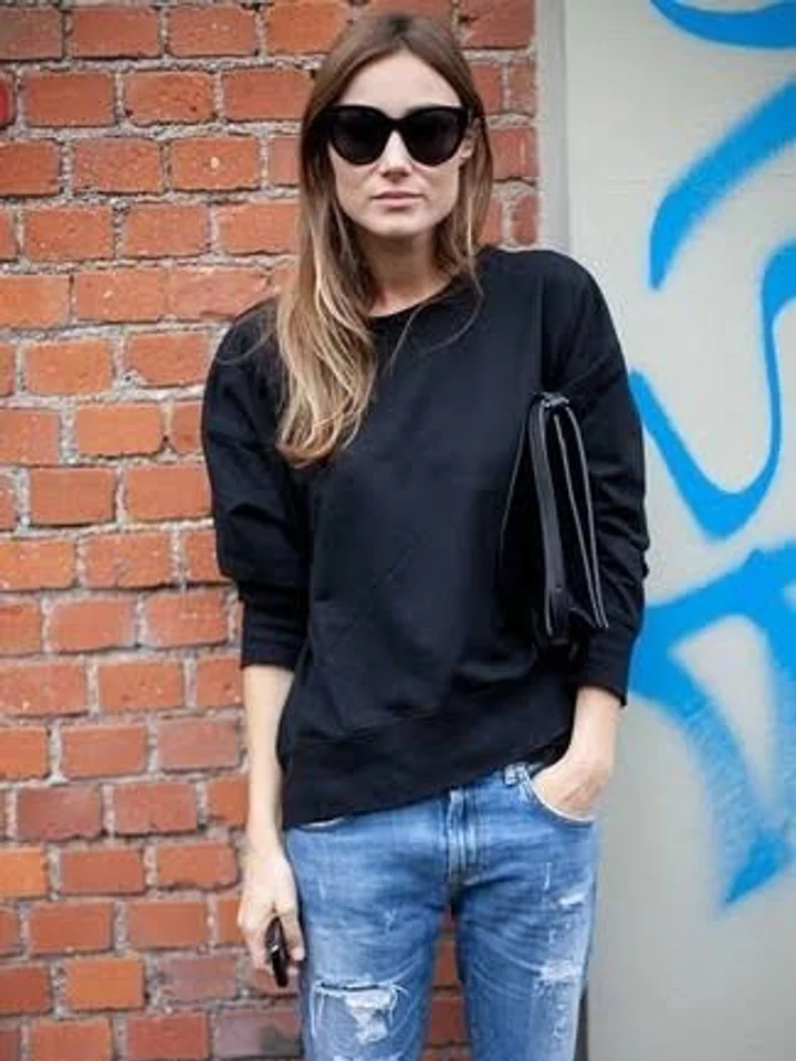 A customer in a plain black crewneck sweatshirt and distressed jeans, wearing sunglasses against a brick wall