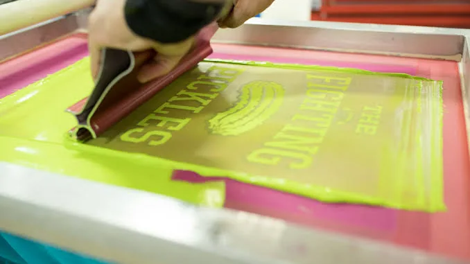 A printer pulling a squeegee across a screen of neon green ink on a screen-printing press