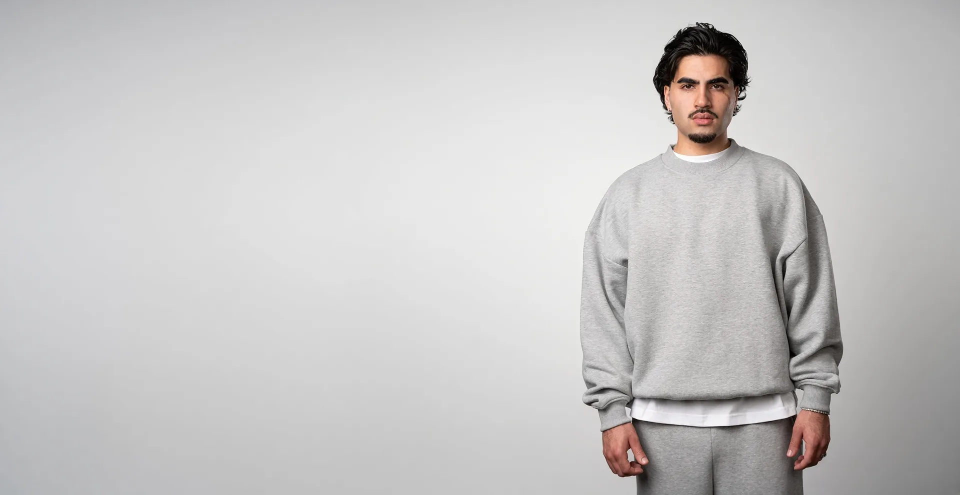 A model in an oversized plain light-grey crewneck sweatshirt and matching joggers against a light grey studio backdrop