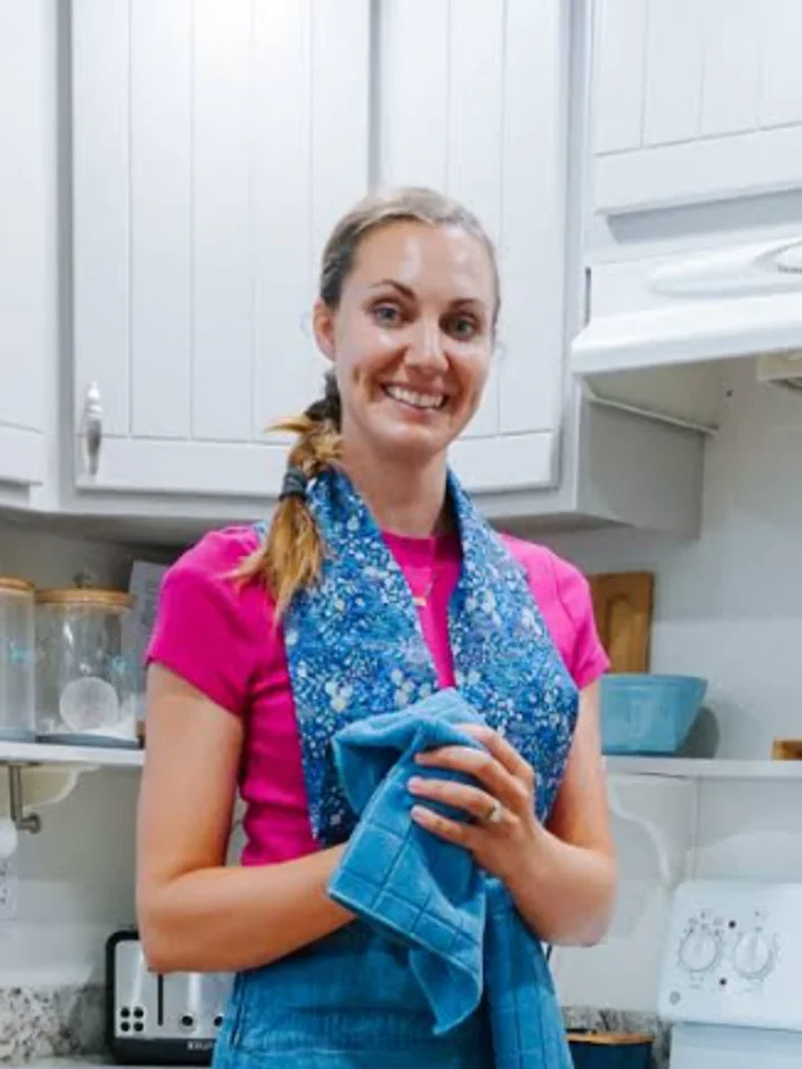 In a pink tee and denim apron, a woman holds a folded blue print towel in her kitchen