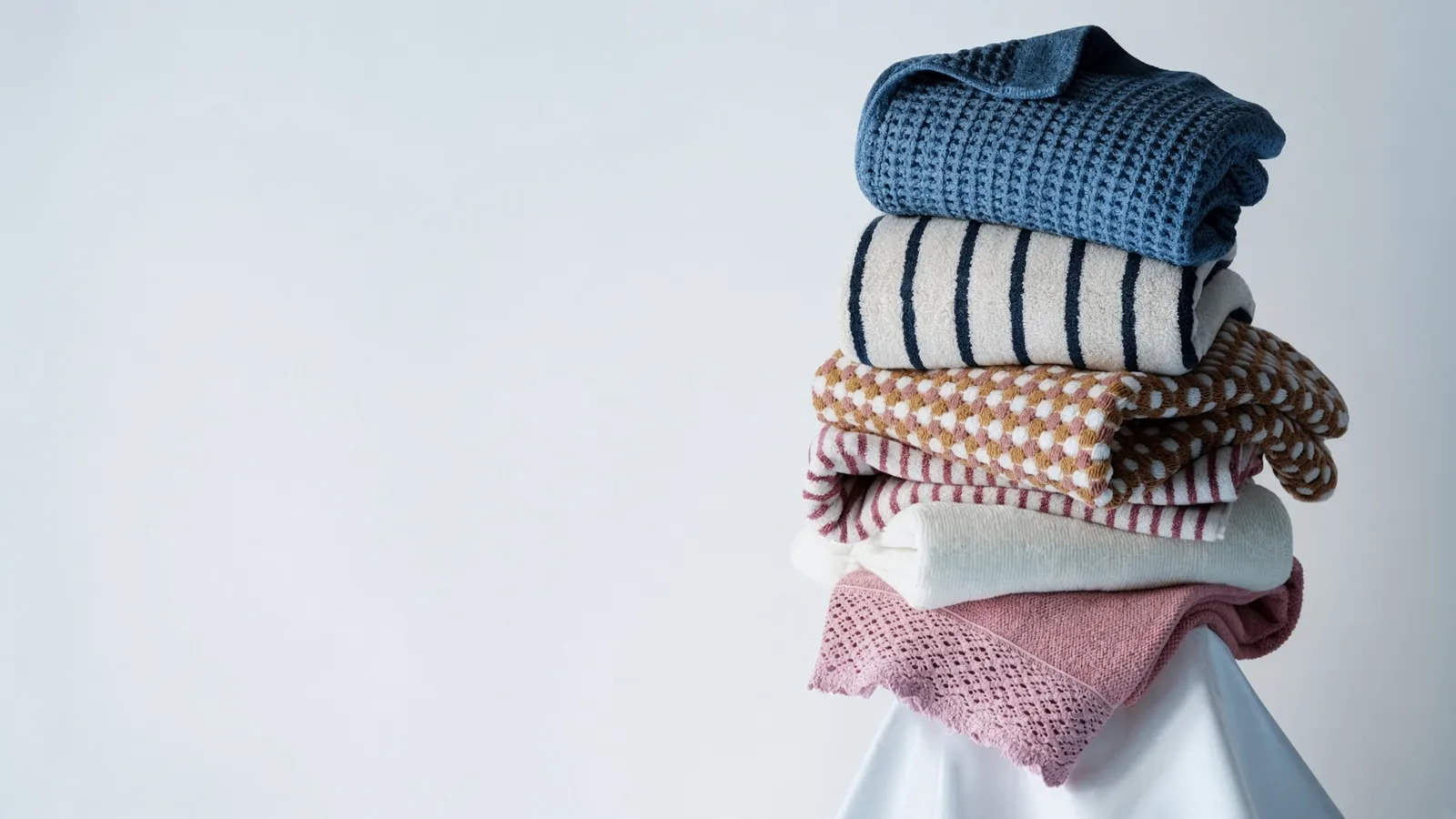 Towels in blue waffle, navy stripe, mustard check and pink lace, stacked on a pale ground